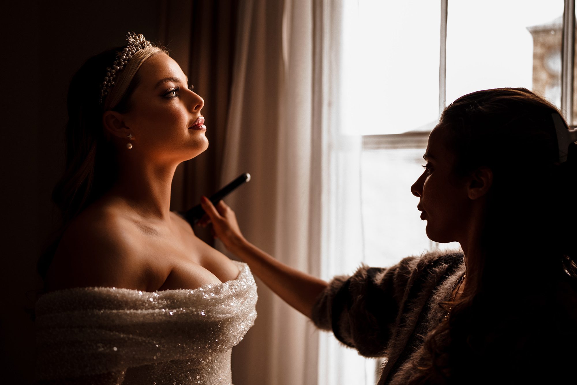 makeup artist appling make up to bride next to window